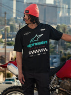 Alpinestars Aplomb Oversized CSF Tee, t-shirt a maniche corte, nero, con grande logo Astars sfumato verde acqua e bianco e grafica con bandiera a scacchi, indossata con un berretto corallo, in piedi accanto a una motocicletta rossa, streetwear casual lifestyle