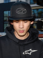 Alpinestars Contender Trucker Hat, berretto lifestyle nero con retro in rete di poliestere, corona strutturata con patch ricamata del logo Astars, indossato da un uomo con felpa nera Alpinestars, streetwear casual ispirato al motorsport