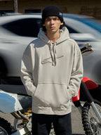Alpinestars Emergent Oversized Hoodie in Off White, felpa oversize con logo Astars ricamato in 3D, tasca a marsupio e cappuccio con cordino, indossata da un uomo con un berretto nero accanto a una moto, stile casual lifestyle motorsport streetwear