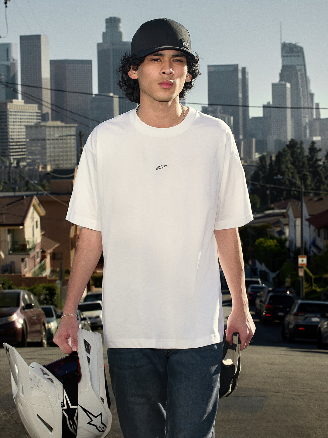 Alpinestars Focused Oversized CSF Tee, t-shirt a maniche corte, bianco, con piccolo logo Astars nero sul petto, scatto lifestyle di una persona con casco da motocross e skyline cittadino sullo sfondo, streetwear casual motorsport