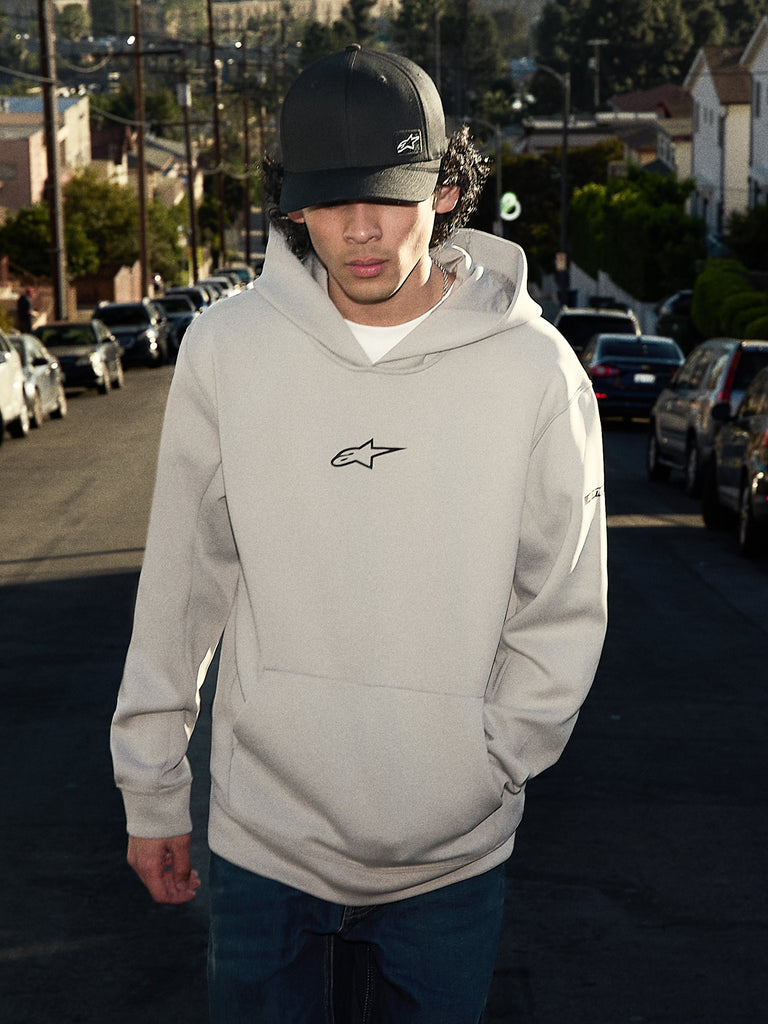 Alpinestars Insight Hoodie, abbigliamento casual lifestyle streetwear, grigio cemento con logo Astars nero sul petto, modello pullover con tasca a marsupio, indossata con un cappellino nero in una strada residenziale, design moderno ispirato al motorsport