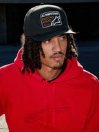 Uomo che indossa il cappello Alpinestars Intuitive Snapback nero, con corona strutturata e patch con logo ricamato 'Racing Ahead', abbinato a una felpa con cappuccio rosso acceso, stile streetwear casual lifestyle.
