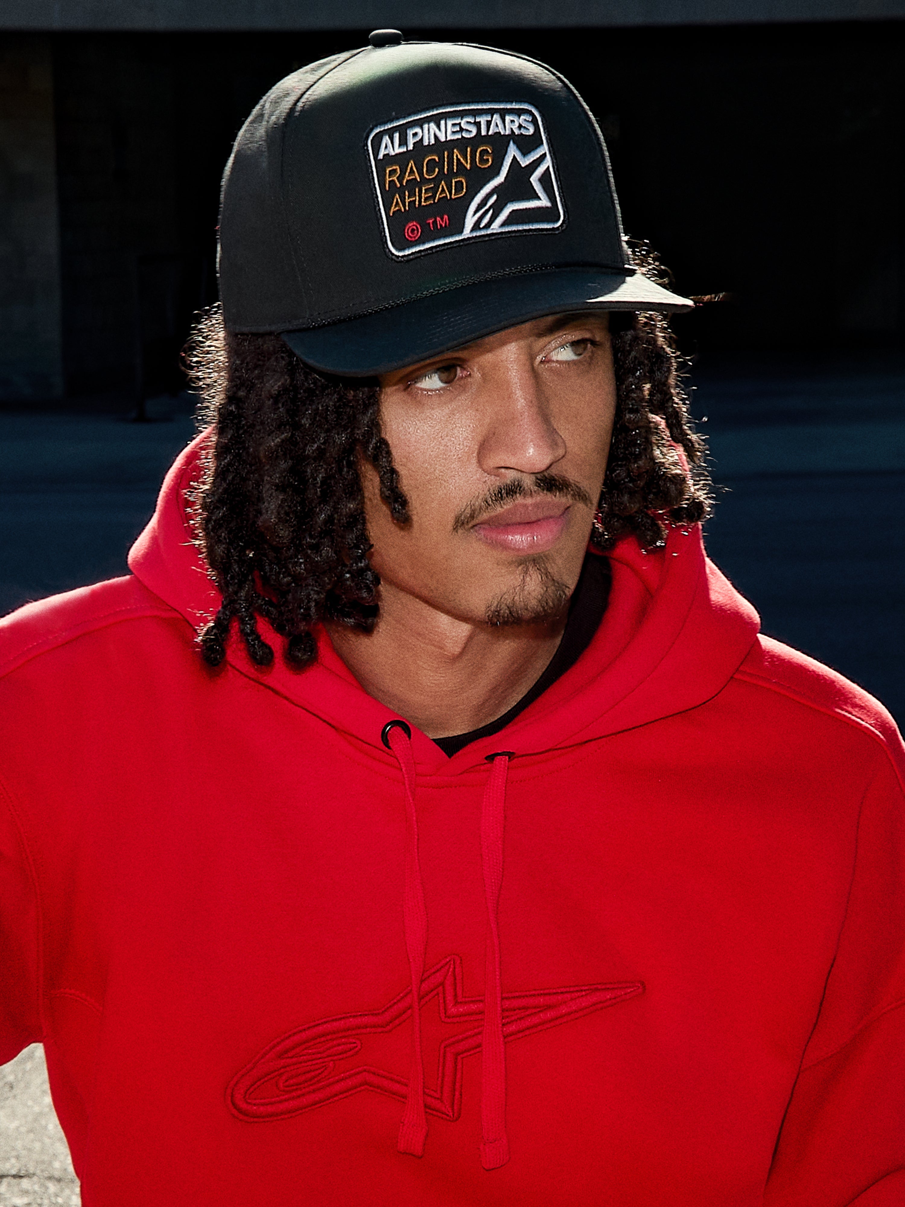 Uomo che indossa il cappello Alpinestars Intuitive Snapback nero, con corona strutturata e patch con logo ricamato 'Racing Ahead', abbinato a una felpa con cappuccio rosso acceso, stile streetwear casual lifestyle.