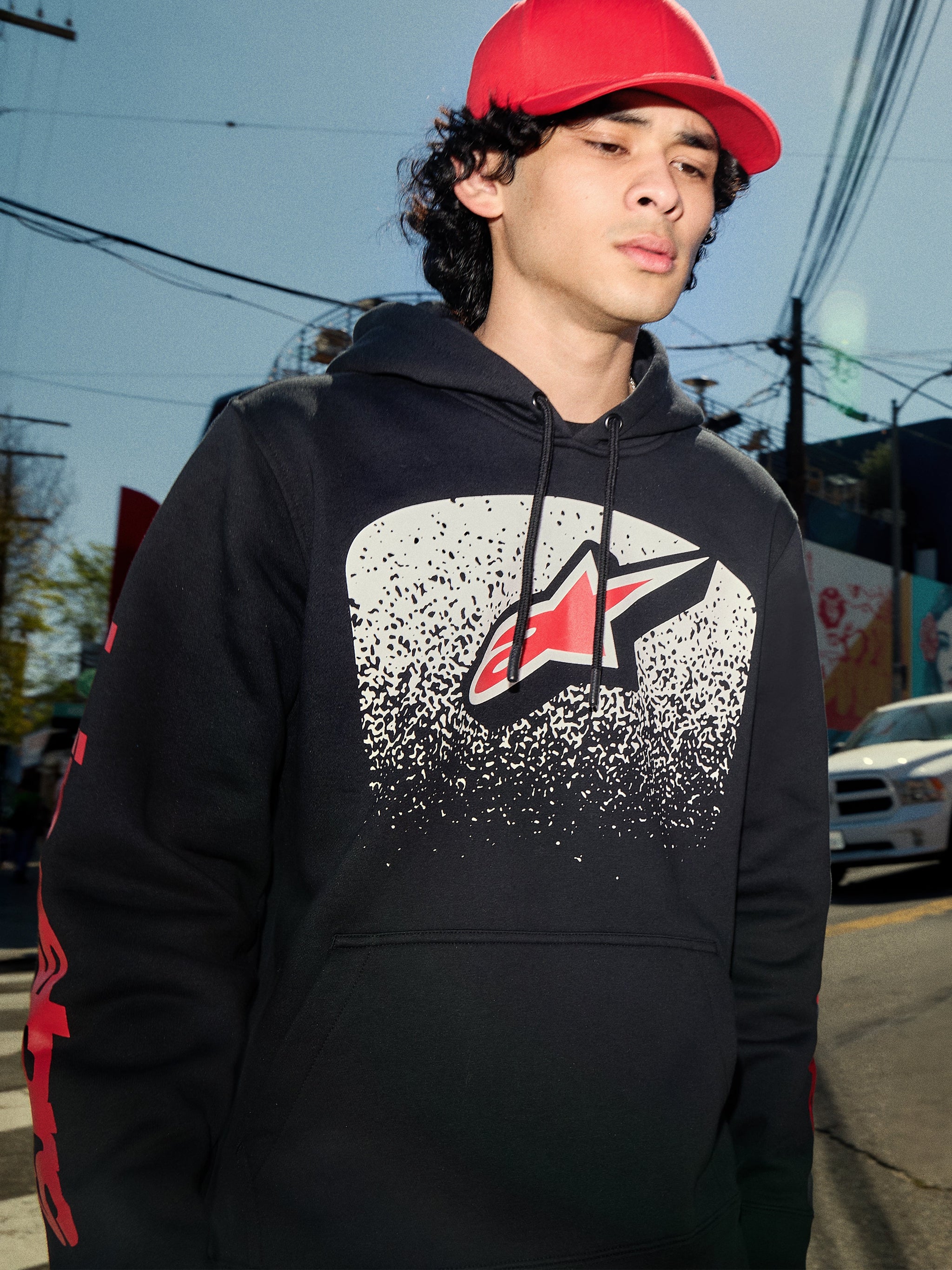 Alpinestars Obsessed Hoodie, felpa in pile nera, grafica sul petto con gradazione MX bianca e logo a stella rossa, loghi rossi sulle maniche, streetwear casual di ispirazione motorsport in un contesto urbano