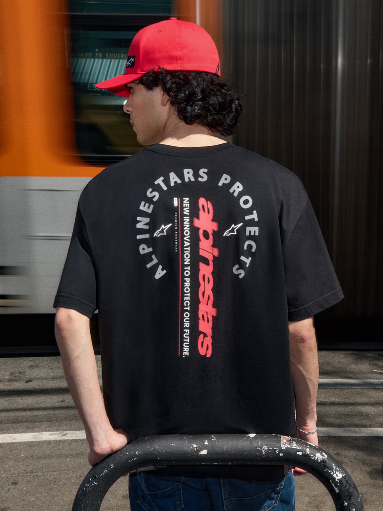 Alpinestars Overhang Oversized CSF Tee, t-shirt nera a maniche corte, vista posteriore con marchio testuale rosso verticale e grafica circolare Alpinestars Protects, indossata con un cappellino rosso, streetwear motorsport casual