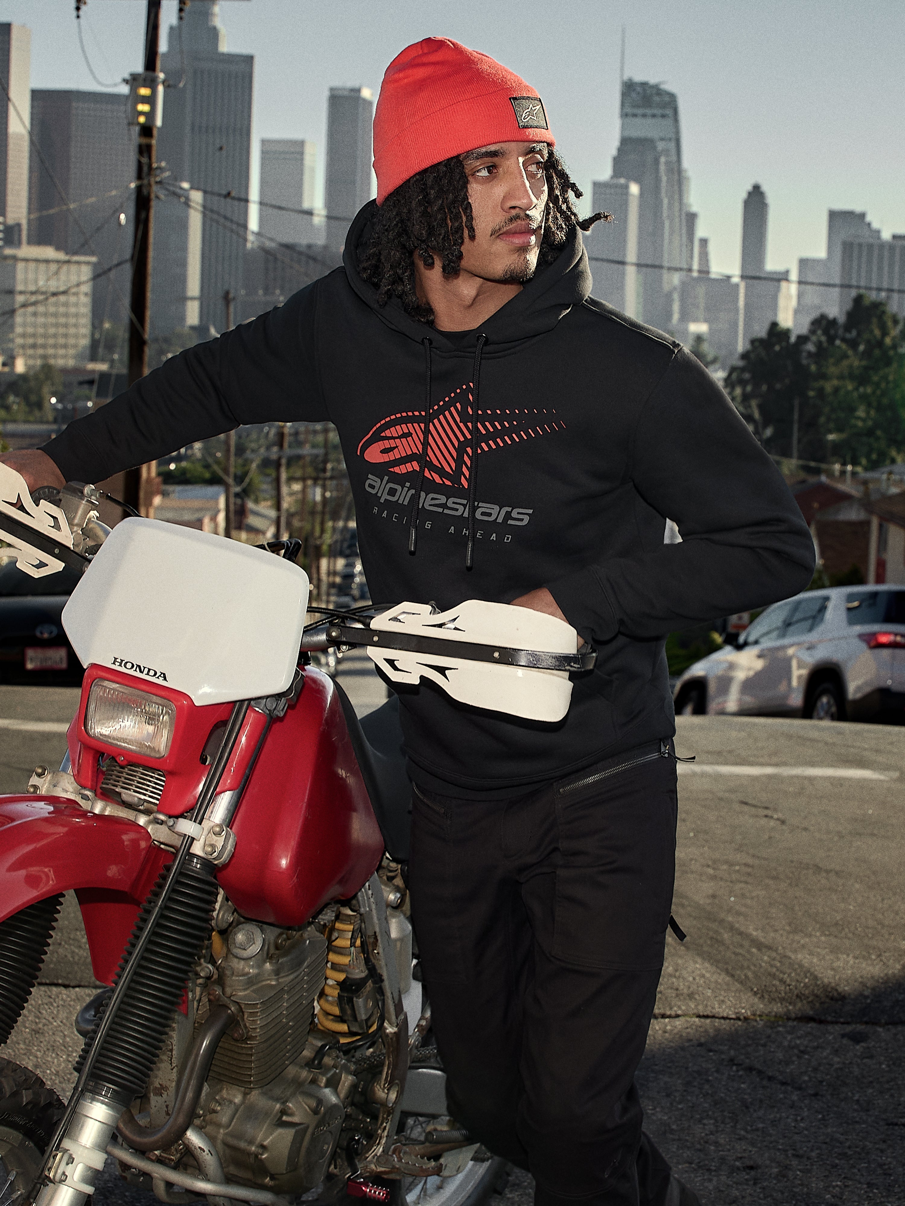 Alpinestars Pulse Hoodie, felpa con cappuccio, nera con logo Astars Combo rosso e grigio, indossata da un uomo con berretto arancione accanto a una motocicletta Honda rossa in un contesto urbano, streetwear casual motorsport