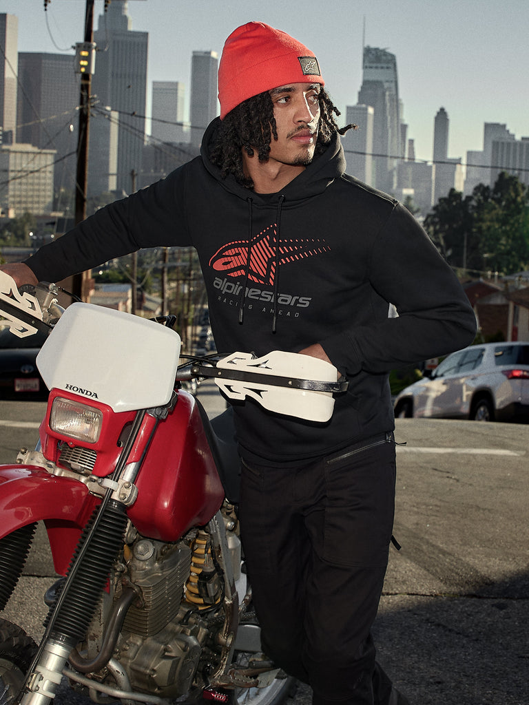 Alpinestars Pulse Hoodie, felpa con cappuccio, nera con logo Astars Combo rosso e grigio, indossata da un uomo con berretto arancione accanto a una motocicletta Honda rossa in un contesto urbano, streetwear casual motorsport
