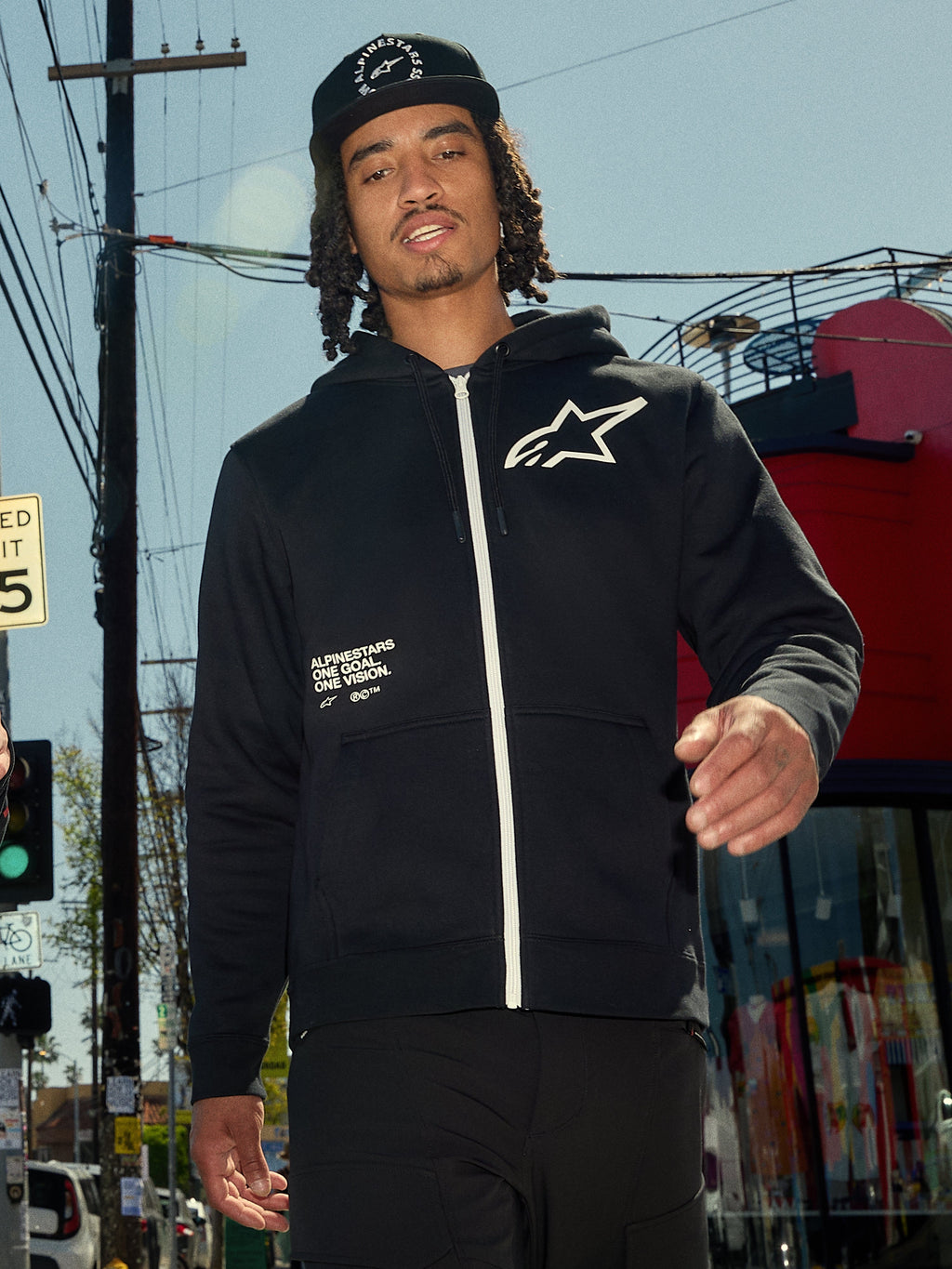 Felpa con cappuccio Alpinestars Transverse, felpa in pile nera con zip frontale, zip bianca e logo Astars bianco sul petto, stampa con testo 'One Goal One Vision', stile streetwear casual, indossata da un uomo in un ambiente urbano all'aperto