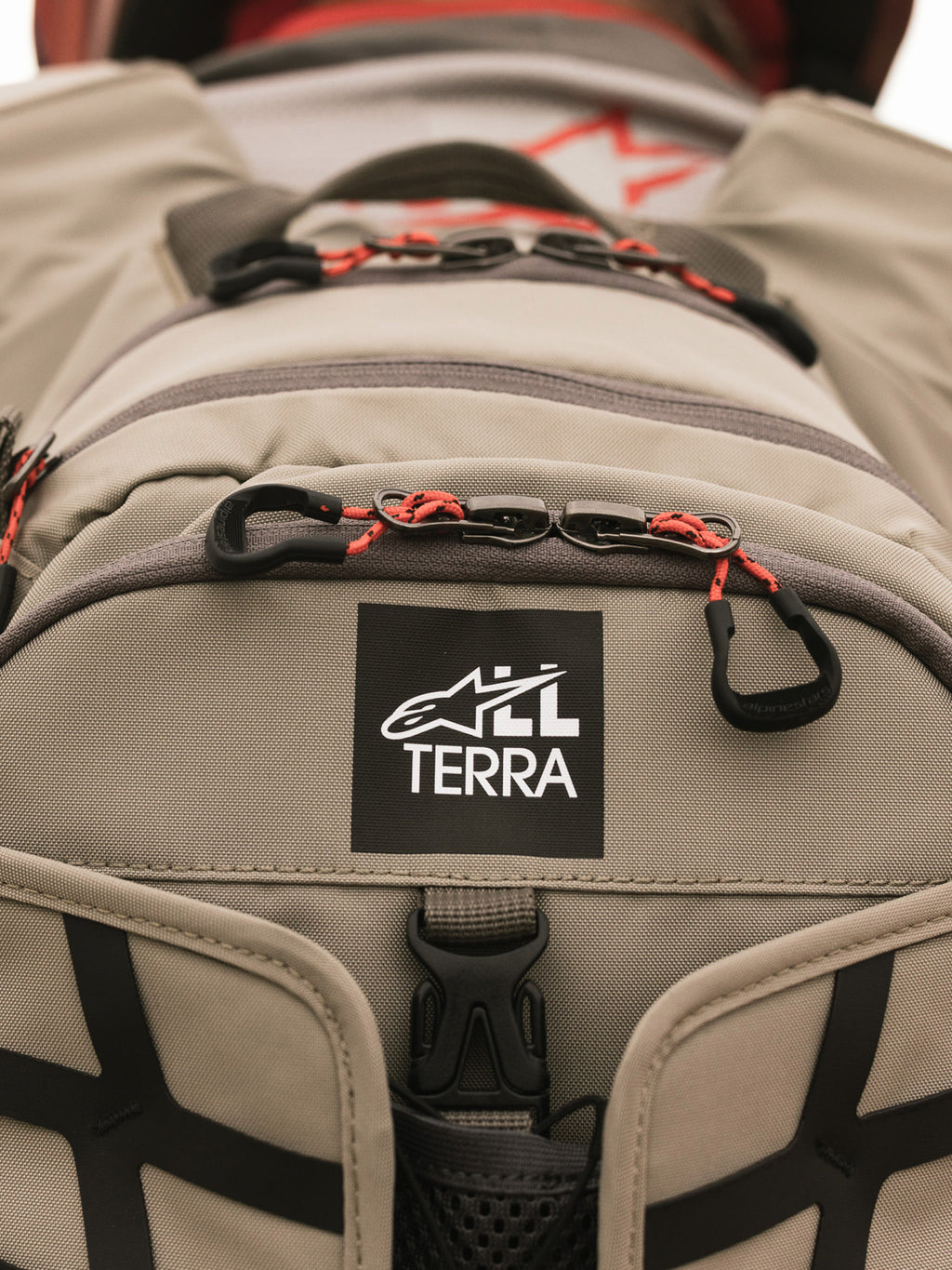 Alpinestars AT-15 Back Pack, Aluminum Walnut, vista ravvicinata della toppa con logo All Terra su tessuto tecnico color sabbia, con tiretti delle cerniere in cordino arancione e dettagli di costruzione robusti