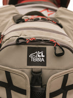 Alpinestars AT-15 Back Pack, Aluminum Walnut, vista ravvicinata della toppa con logo All Terra su tessuto tecnico color sabbia, con tiretti delle cerniere in cordino arancione e dettagli di costruzione robusti