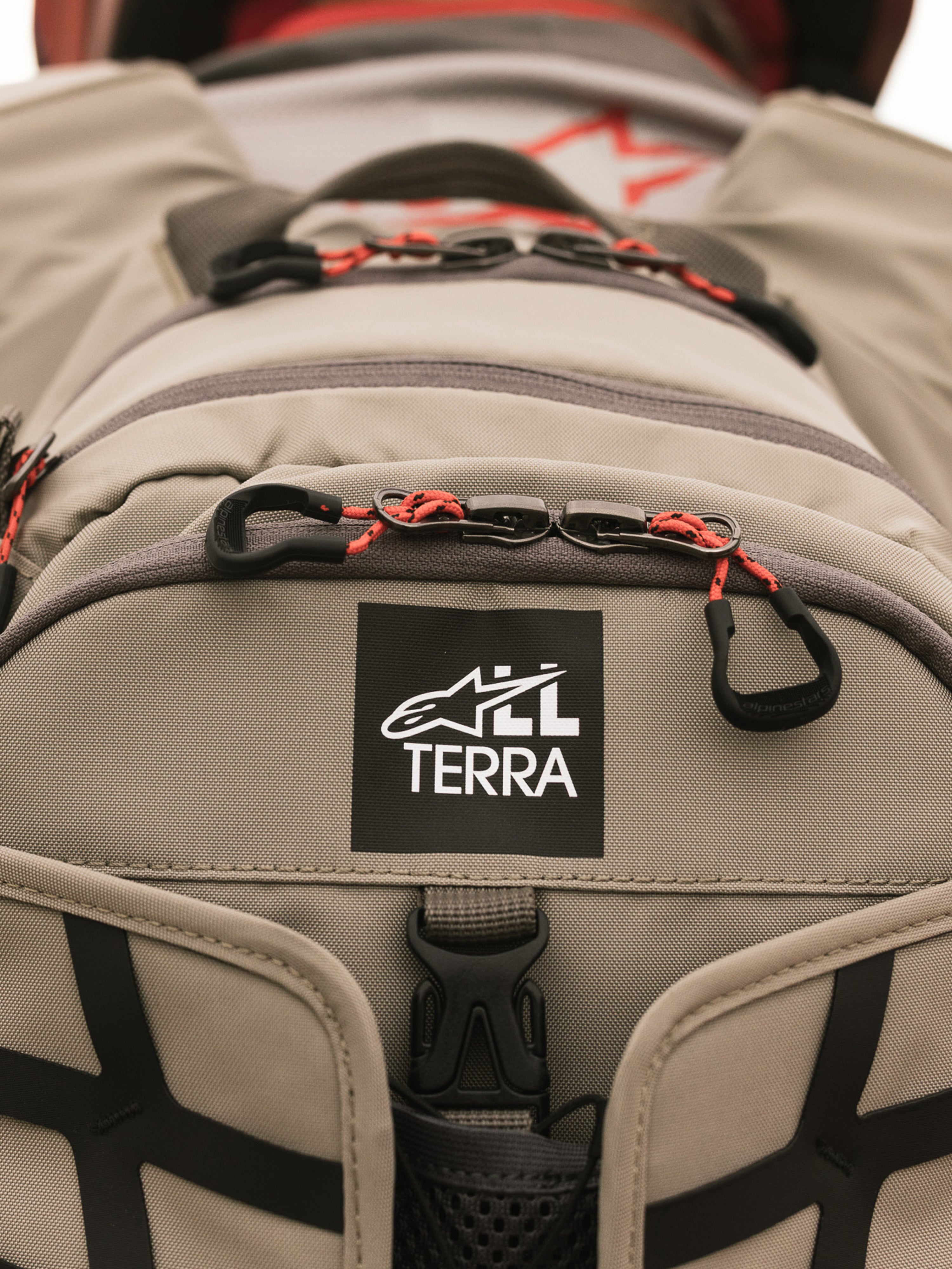 Alpinestars AT-15 Back Pack, Aluminum Walnut, vista ravvicinata della toppa con logo All Terra su tessuto tecnico color sabbia, con tiretti delle cerniere in cordino arancione e dettagli di costruzione robusti