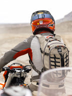 Zaino Alpinestars AT-15 in colore Aluminum Walnut, indossato da un motociclista adventure in un paesaggio desertico. Presenta una robusta struttura in tessuto beige con cinghie nere, dettagli delle cerniere arancioni e marchio 'All Terra'. Abbinato a un casco rosso e arancione e abbigliamento grigio per touring fuoristrada.