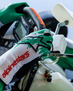 Guanti Alpinestars Tech-1 K V3 in bianco, verde e rosso, mani del pilota che impugnano un volante da kart, design con grafica verde sublimata, logo a stella nero e scritta Alpinestars rossa sul polsino, attrezzatura professionale da kart