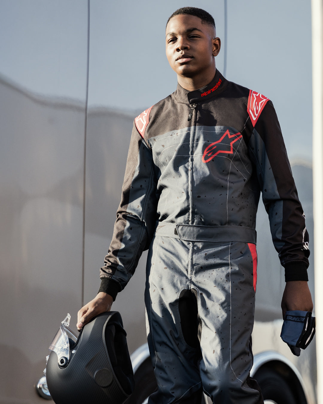 Pilota di kart con la tuta Alpinestars KMX-9 V3 Graphic 6 in nero antracite e rosso, con casco nero opaco e guanti, loghi Alpinestars rossi e design grafico a schizzi, ambientazione motorsport all'aperto.