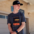 Alpinestars Galun CSF Tee, t-shirt a maniche corte, grigio, grafica frontale con lattina di gas vintage, marchio 'astars' e numeri romani 'MCMLXIII', indossata da un uomo con cappellino nero con logo, stile casual motorsport