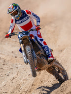 Pilota di motocross in azione su una pista sterrata, indossa la maglia Limited Edition Techstar Fortitude in rosso, bianco e blu, coordinata con pantaloni da gara e stivali abbinati, con logo Alpinestars in evidenza, abbigliamento professionale per gare off-road