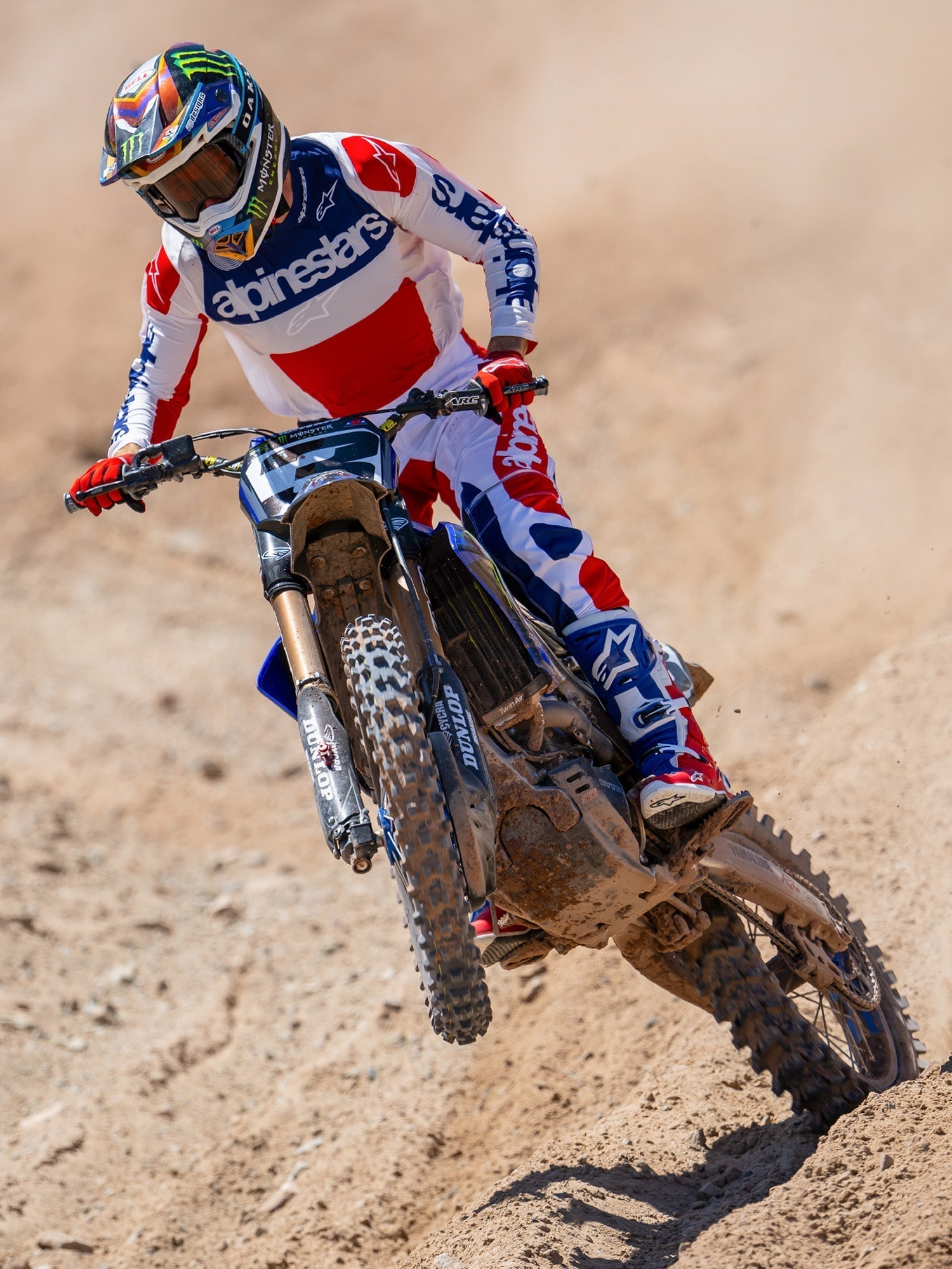 Alpinestars Limited Edition Techstar Fortitude Pants, pantaloni da motocross, rosso brillante, bianco e blu, indossati da un pilota in azione su una pista sterrata, con maglia e stivali coordinati, equipaggiamento da gara off-road ad alte prestazioni.