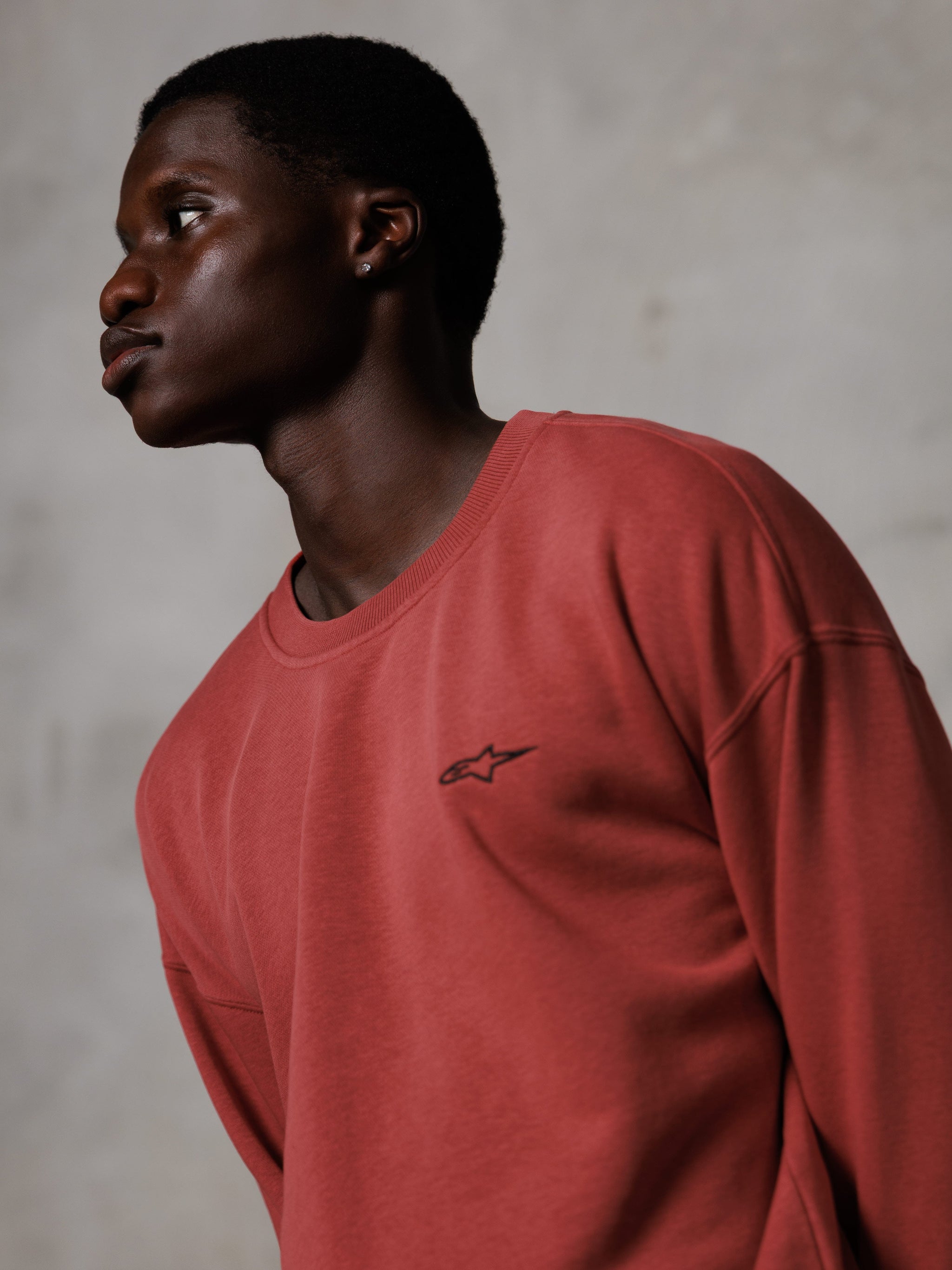 Alpinestars Essentials Ageless Crew, felpa girocollo rosso mattone, indossata da un modello di profilo, piccolo logo a stella nero sul petto, vestibilità rilassata, streetwear casual in pile premium