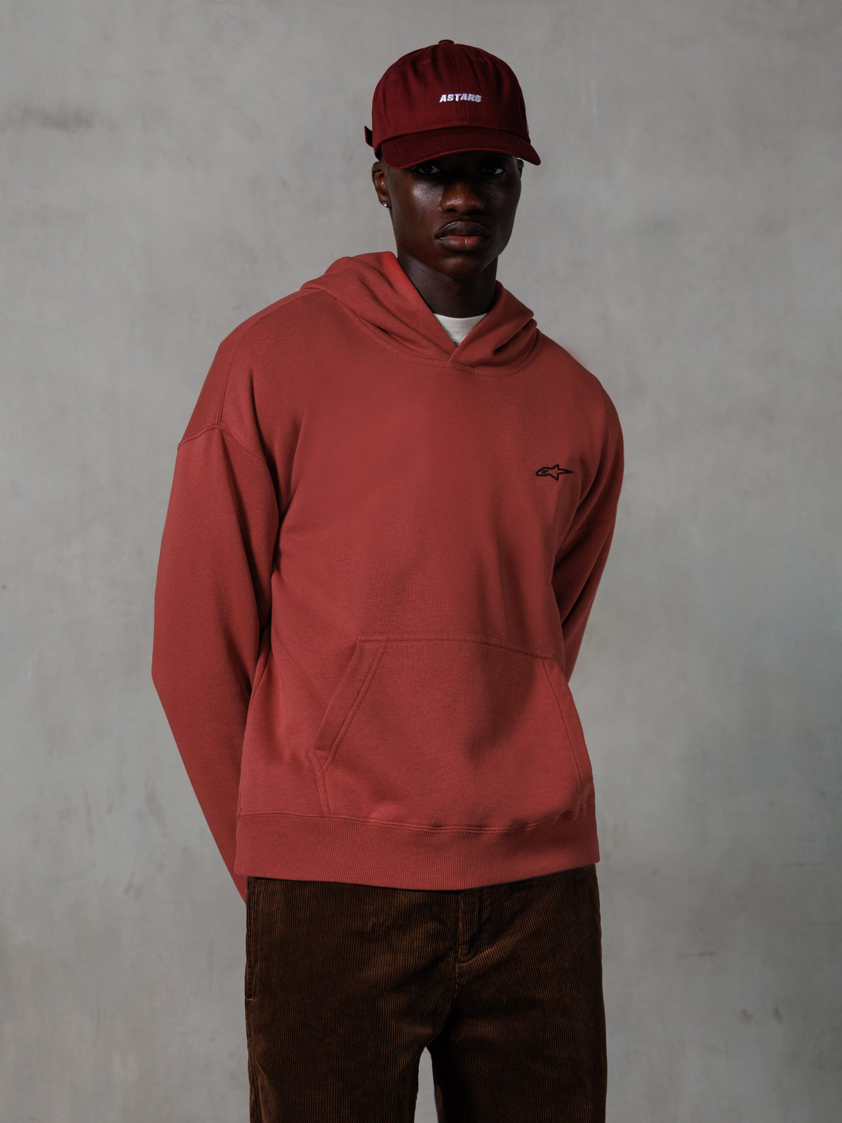 Felpa con cappuccio Alpinestars Essentials Ageless, streetwear casual, rosso mattone, piccolo logo nero sul petto, tasca a marsupio frontale, indossata con cappellino ASTARS rosso coordinato e pantaloni di velluto a coste marroni, design pulito e minimalista