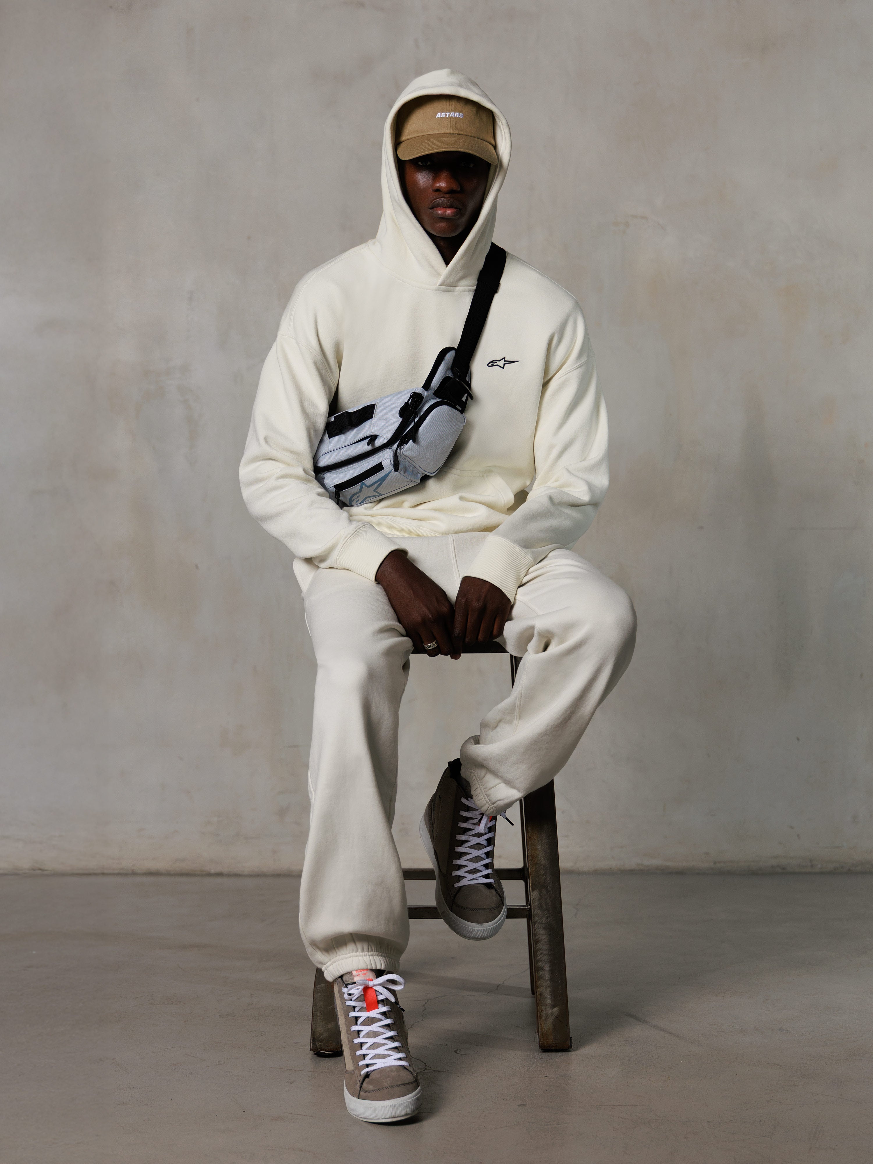 Alpinestars Essentials Ageless Hoodie, Off-White, indossata da un modello seduto su uno sgabello con pantaloni jogger in pile coordinati, cappellino ASTARS beige e borsa a tracolla, streetwear casual ispirato al motorsport