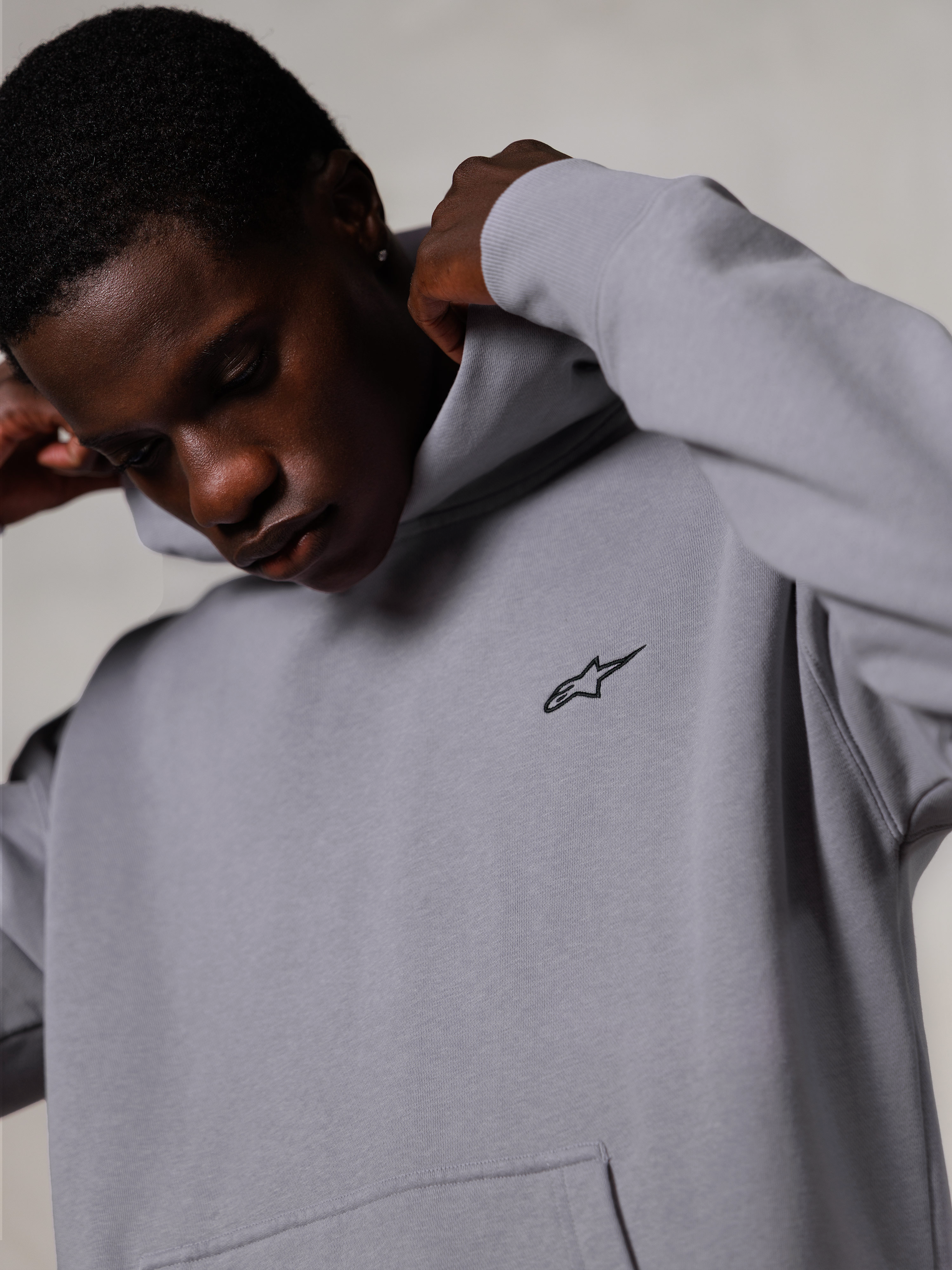 Alpinestars Essentials Ageless Hoodie, Silver, primo piano di un uomo che sistema il cappuccio della felpa grigia, piccolo logo Alpinestars nero sul petto, abbigliamento casual premium ispirato al motorsport