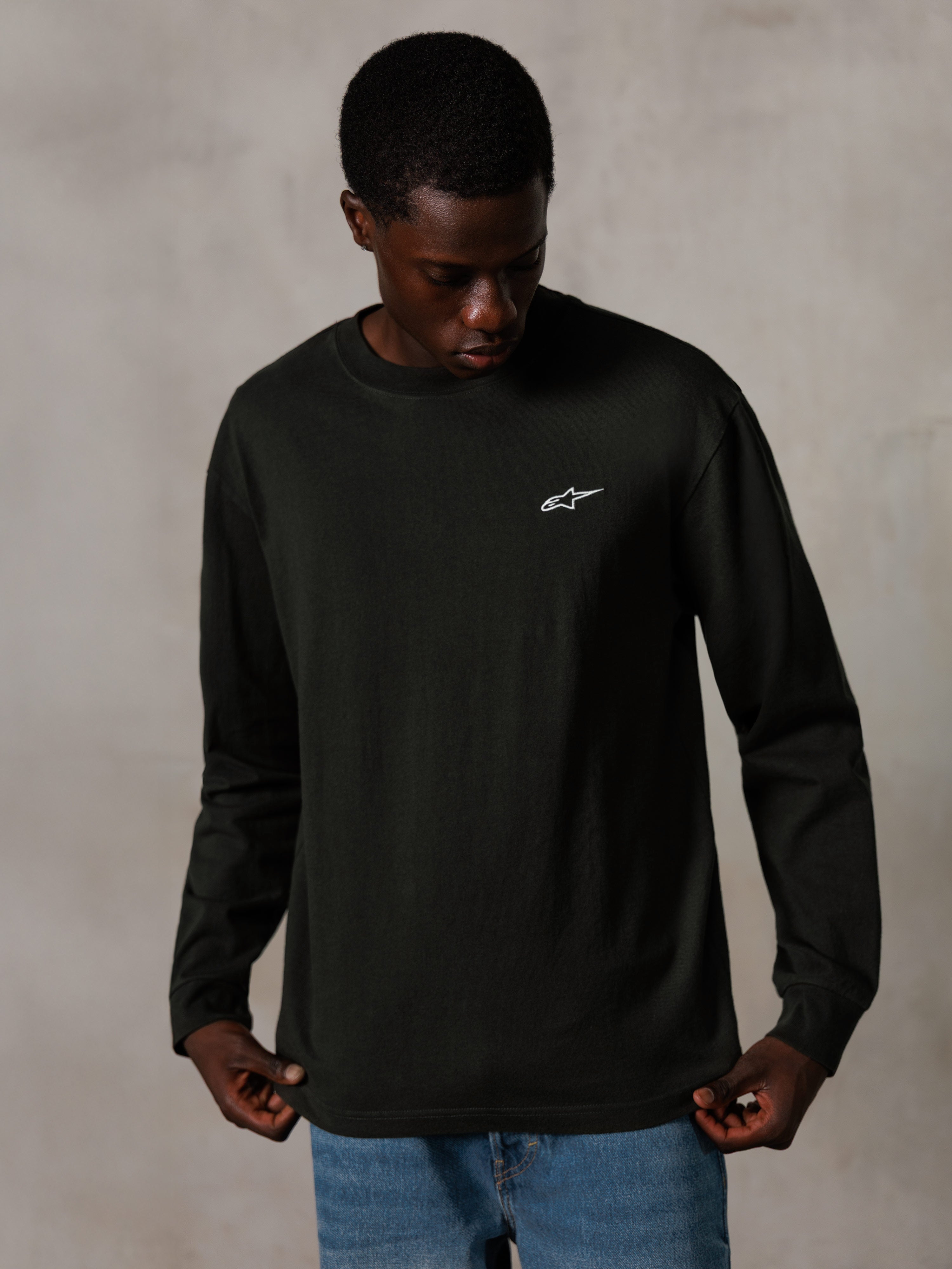 Alpinestars Essentials Ageless LS Tee, t-shirt a maniche lunghe, Off Black, con un piccolo logo bianco sul petto, indossata da un modello con jeans blu, stile streetwear casual