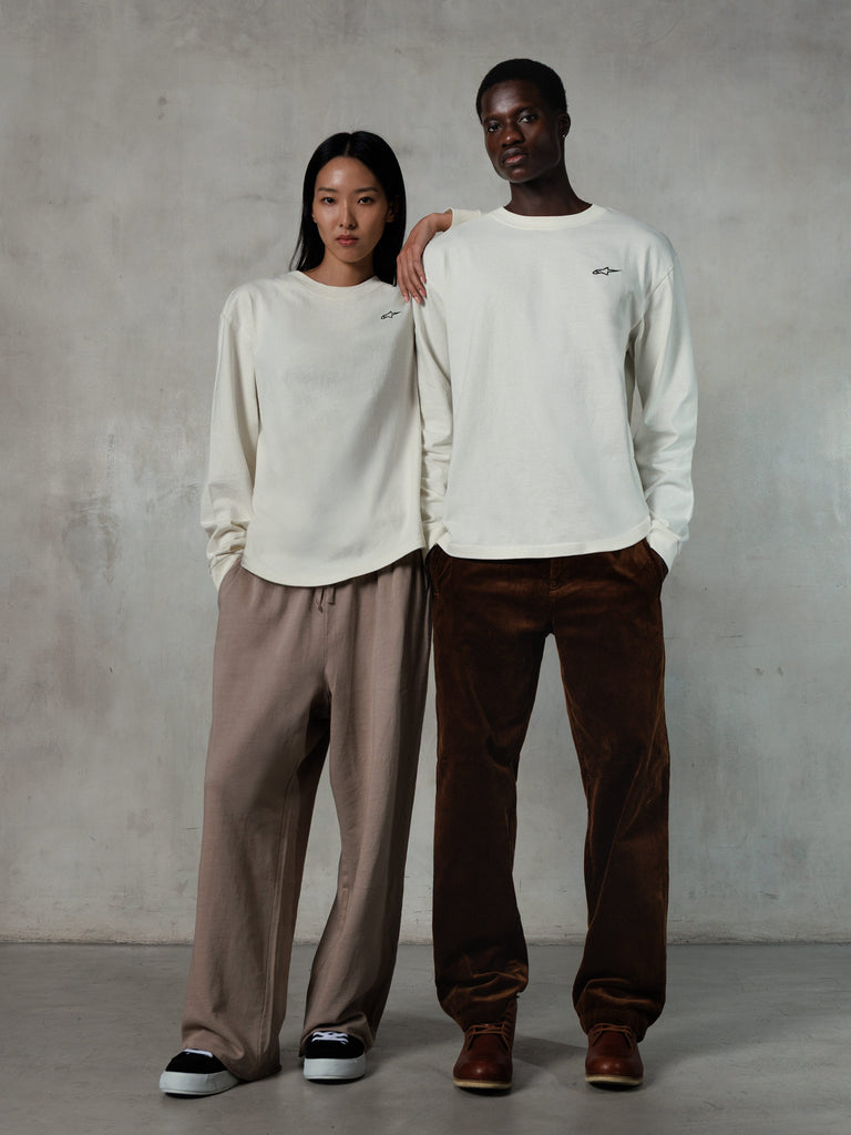 Alpinestars Essentials Ageless LS Tee, t-shirt a maniche lunghe, bianco sporco, indossata da modelli uomo e donna, piccolo logo Alpinestars nero sul petto, vestibilità rilassata, abbinata a pantaloni della tuta beige e pantaloni in velluto a coste marroni, streetwear casual ispirato al motorsport