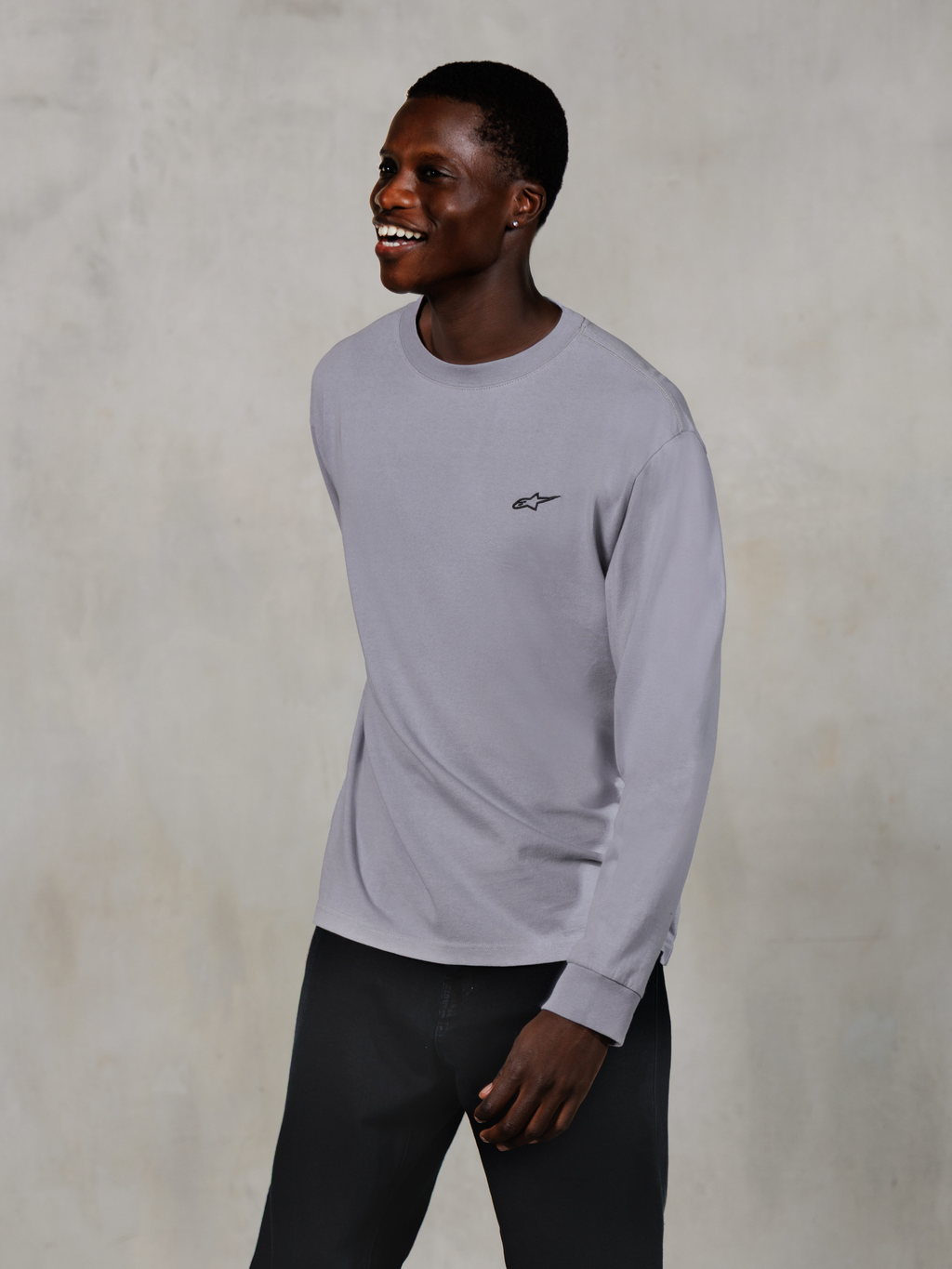 Alpinestars Essentials Ageless LS Tee, maglietta a maniche lunghe, Silver, piccolo logo nero sul petto, indossata da un modello sorridente con pantaloni scuri, vestibilità rilassata contemporanea, abbigliamento casual motorsport lifestyle