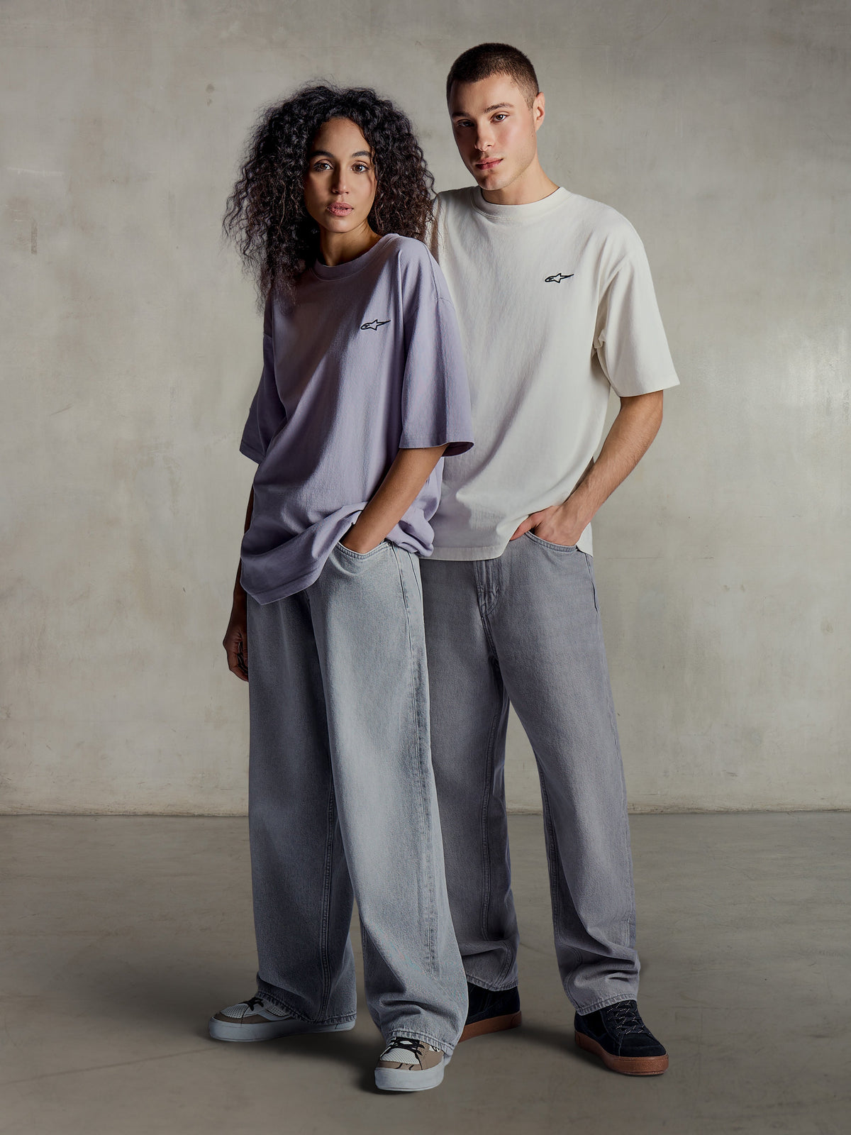Alpinestars Essentials Ageless SS Tee, vista lifestyle di due modelli che indossano magliette a maniche corte in viola chiaro e argento, cotone organico con vestibilità rilassata e un sottile logo sul petto, abbinate a jeans larghi per uno stile streetwear casual.