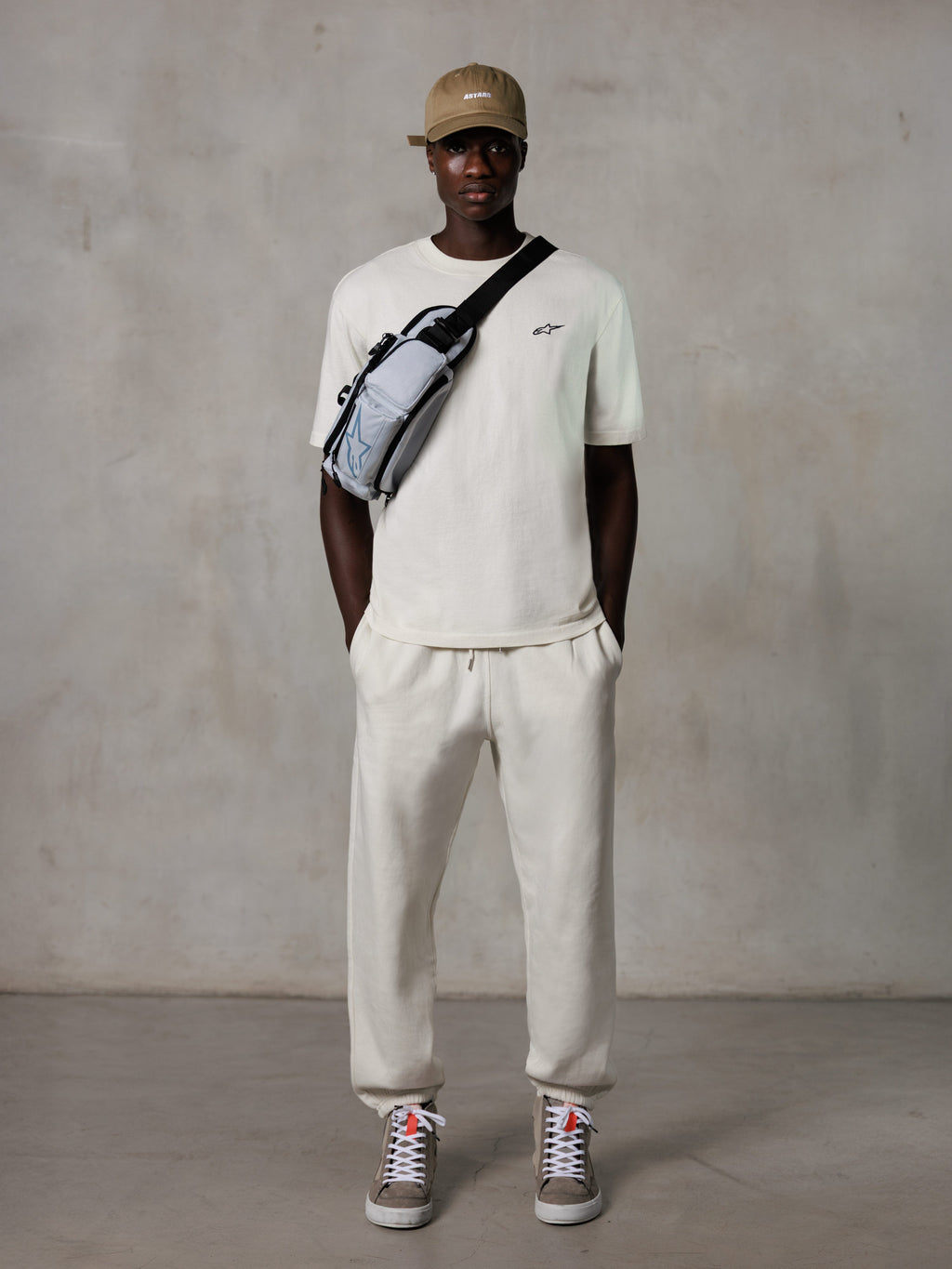 Alpinestars Essentials Ageless SS Tee, t-shirt a maniche corte, off-white, con un piccolo logo nero sul petto, indossata con joggers coordinati, cappellino da baseball color sabbia e borsa a tracolla grigia, streetwear casual lifestyle