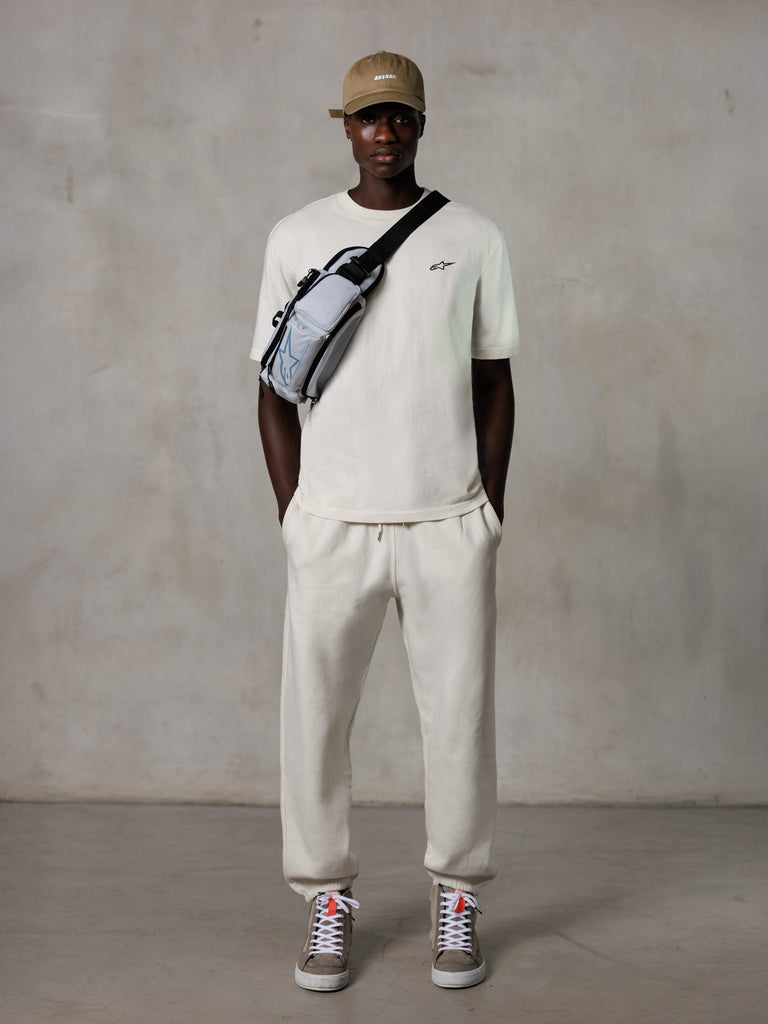 Alpinestars Essentials Ageless SS Tee, t-shirt a maniche corte, off-white, con un piccolo logo nero sul petto, indossata con joggers coordinati, cappellino da baseball color sabbia e borsa a tracolla grigia, streetwear casual lifestyle