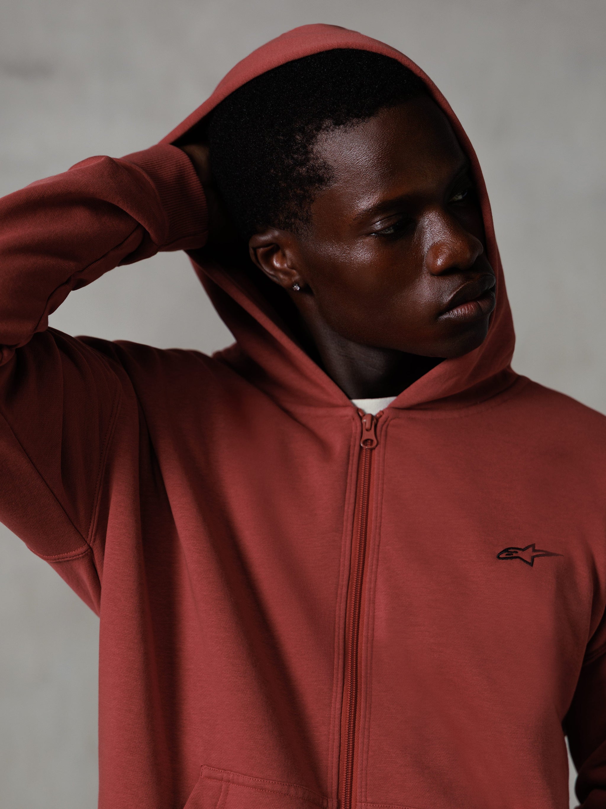 Alpinestars Essentials Ageless Zip Hoodie, rosso mattone, lifestyle streetwear casual, cappuccio alzato con mano dietro la testa, piccolo logo Astars nero sul petto, felpa in pile dalla vestibilità rilassata