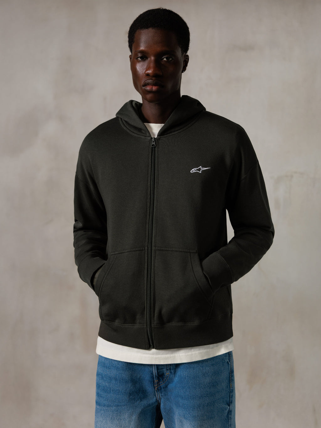 Alpinestars Essentials Ageless Zip Hoodie, Off Black, felpa con cappuccio e zip con un piccolo logo Alpinestars bianco sul petto, vestibilità rilassata, indossata da un modello con jeans blu e una t-shirt bianca, streetwear casual ispirato al motorsport