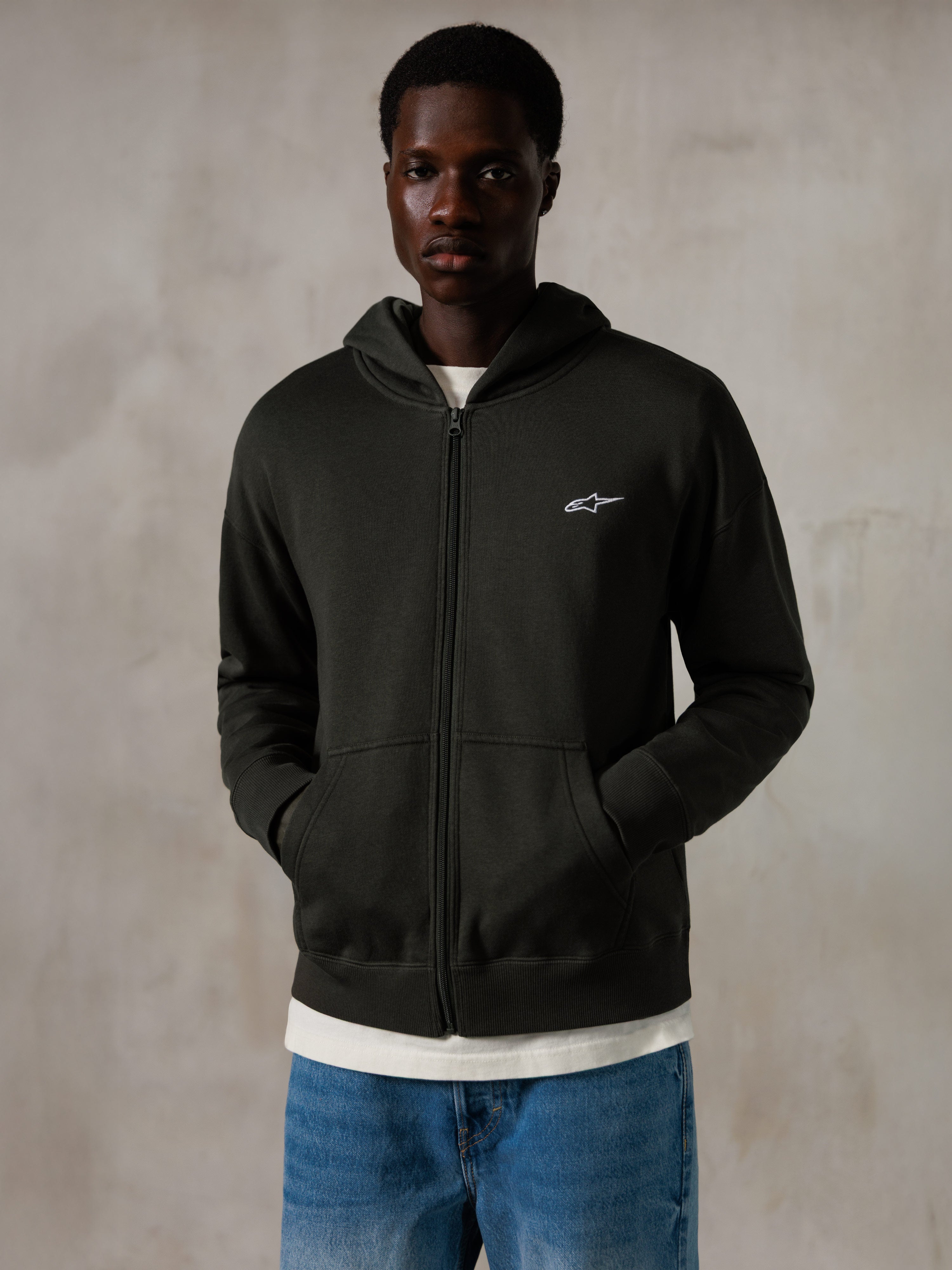 Alpinestars Essentials Ageless Zip Hoodie, Off Black, felpa con cappuccio e zip con un piccolo logo Alpinestars bianco sul petto, vestibilità rilassata, indossata da un modello con jeans blu e una t-shirt bianca, streetwear casual ispirato al motorsport