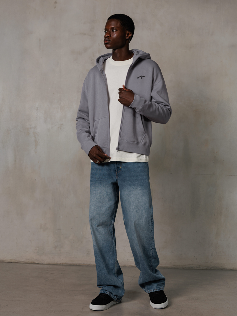 Alpinestars Essentials Ageless Zip Hoodie, Silver, streetwear casual, felpa in pile con cappuccio e zip intera con un piccolo logo nero sul petto, indossata da un modello con t-shirt bianca e jeans blu, vestibilità lifestyle rilassata