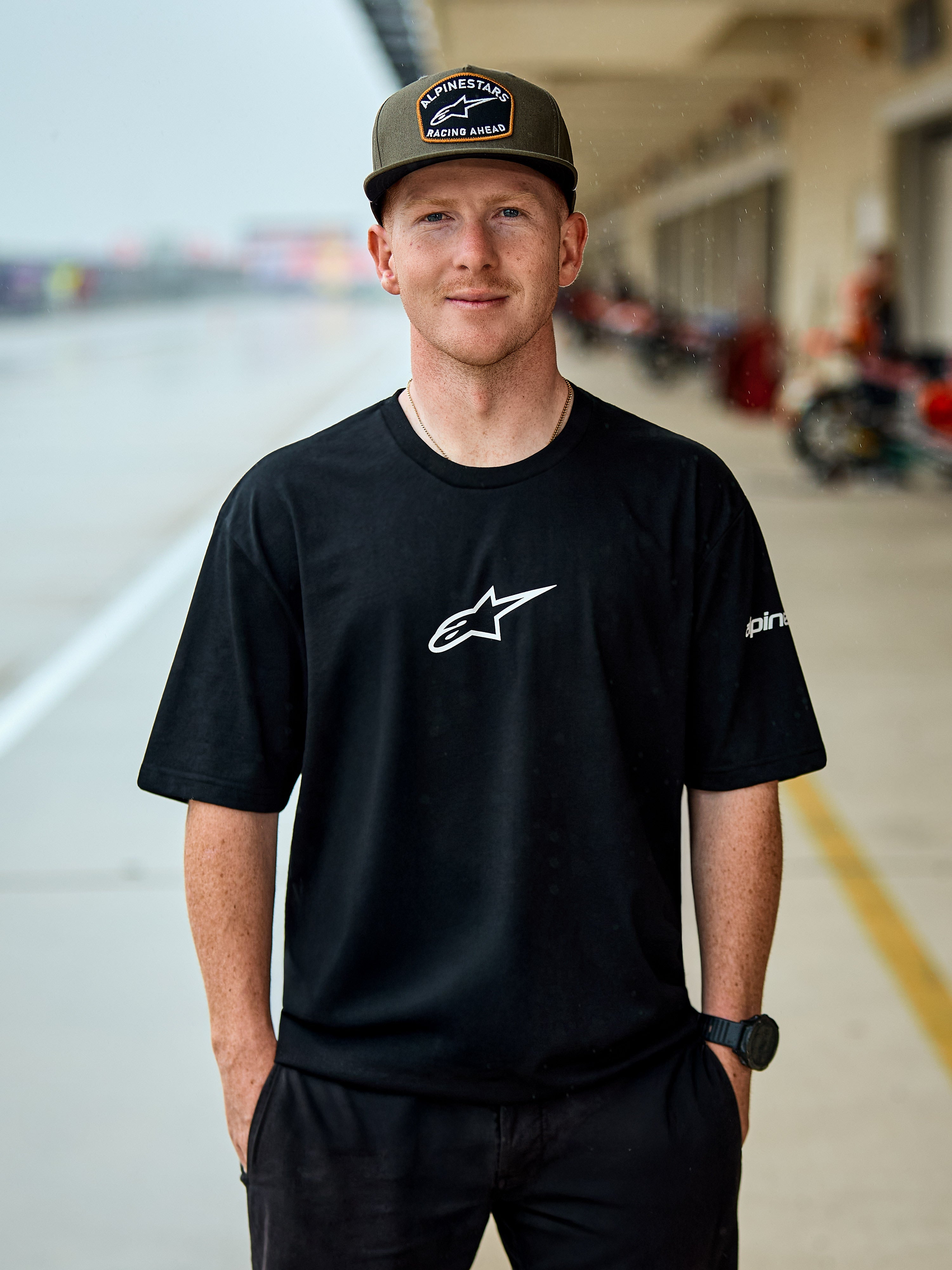 Uomo con t-shirt Alpinestars Frontal CSF, maglietta a maniche corte nera, logo Astars centrale bianco, cappellino Alpinestars verde oliva, streetwear casual ispirato al motorsport