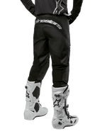 Alpinestars Pantaloni Fluid Graphite 2024, pantaloni da motocross, nero e argento, vista posteriore con logo Alpinestars argento sul carré, indossati con stivali da motocross bianchi e neri, design in tessuto resistente per la guida fuoristrada