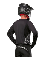 Pilota di motocross visto di spalle con maglia Alpinestars Fluid Graphite 2024 in nero e argento, caratterizzata da un grande logo argento sul lato, casco nero con trama in carbonio e pantaloni neri, abbigliamento professionale per corse fuoristrada