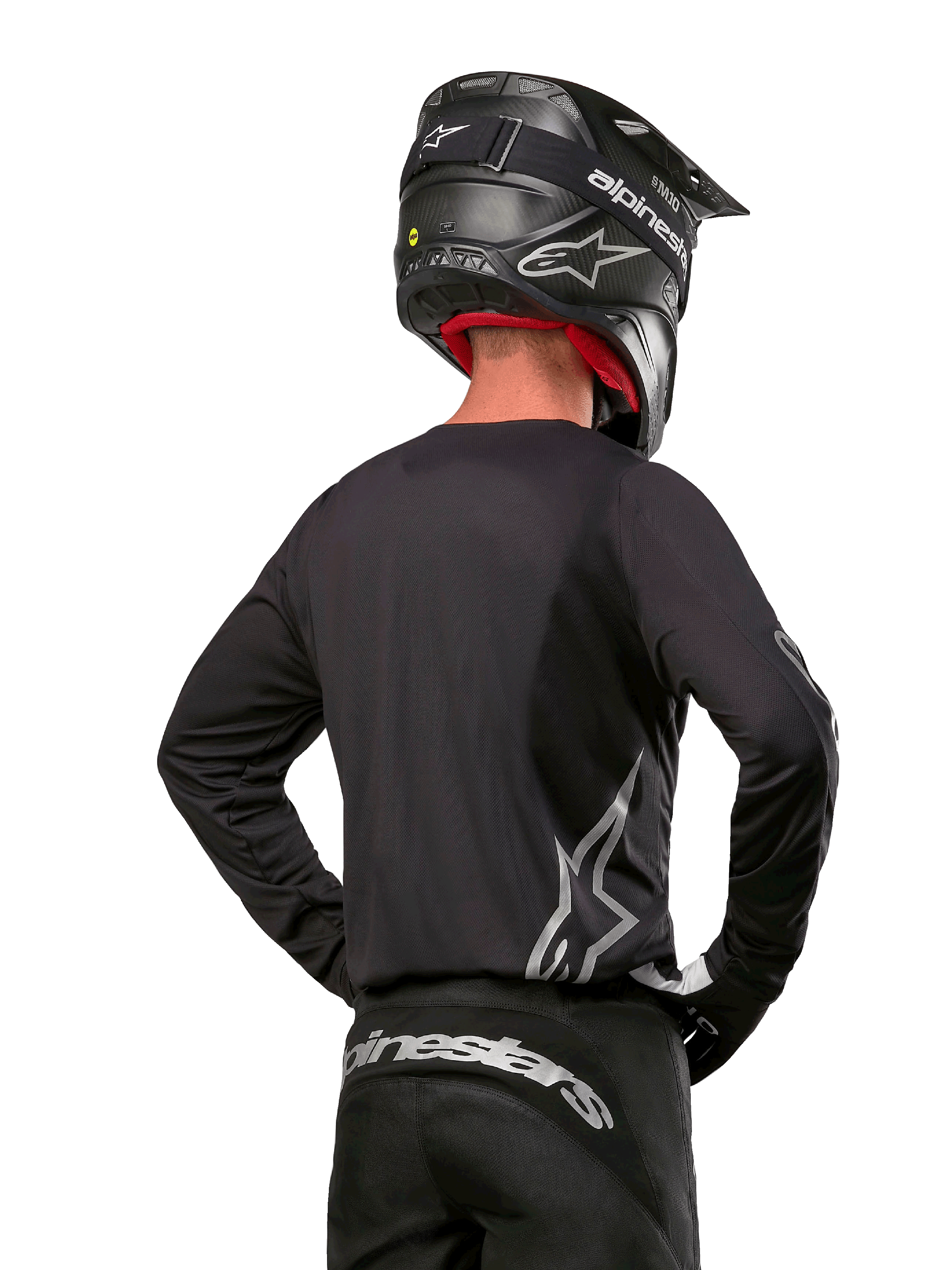 Pilota di motocross visto di spalle con maglia Alpinestars Fluid Graphite 2024 in nero e argento, caratterizzata da un grande logo argento sul lato, casco nero con trama in carbonio e pantaloni neri, abbigliamento professionale per corse fuoristrada