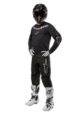 Alpinestars 2024 Fluid Graphite Gear Set, completo motocross nero con loghi grigi e bianchi, maglia e pantaloni abbinati a casco nero coordinato, guanti e stivali bianchi e neri, equipaggiamento protettivo per fuoristrada