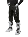 Alpinestars 2024 Fluid Graphite Pants, pantaloni da motocross, nero e argento, con logo Alpinestars argento sulla coscia, mostrati indossati con stivali e guanti da motocross bianchi, progettati per corse motociclistiche fuoristrada