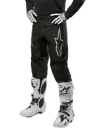Alpinestars 2024 Fluid Graphite Pants, pantaloni da motocross, nero e argento, con logo Alpinestars argento sulla coscia, mostrati indossati con stivali e guanti da motocross bianchi, progettati per corse motociclistiche fuoristrada