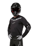Alpinestars 2024 Fluid Graphite Jersey, maglia da motocross nera e argento, vestibilità performance a maniche lunghe, indossata con casco nero e guanti bianchi, con loghi Alpinestars argento, abbigliamento per corse fuoristrada