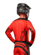 Pilota di motocross visto da dietro con maglia Alpinestars Fluid Lurv 2024 in Hot Orange e nero, loghi Alpinestars su schiena e maniche, abbinata a pantaloni da motocross coordinati e casco nero, progettata per prestazioni off-road.