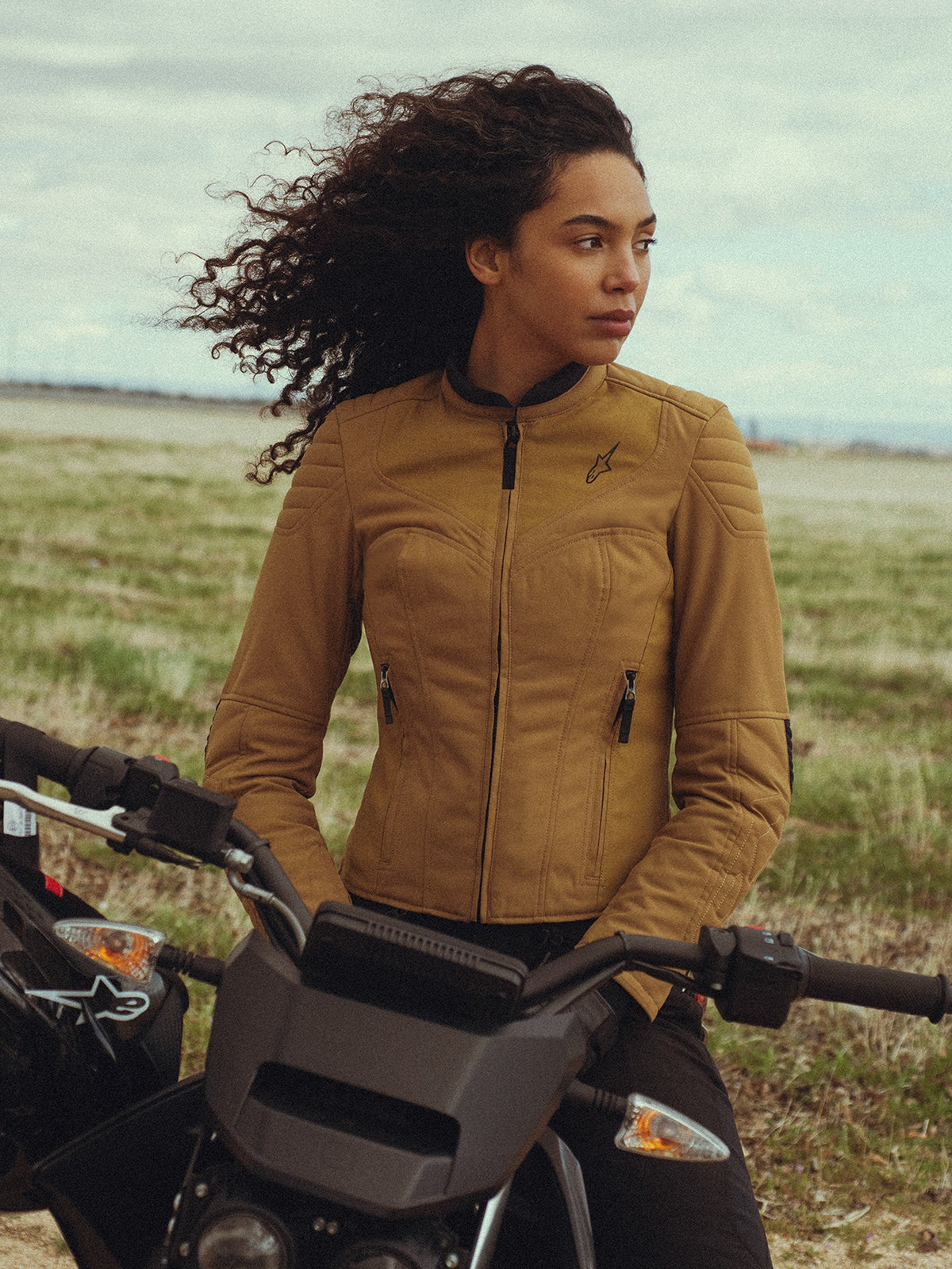 Giacca da donna Alpinestars Isla WR in Utility Brown Light Gold, indossata da una donna seduta su una motocicletta in un campo all'aperto, con dettagli trapuntati sulle spalle, silhouette slim-fit e stile urbano contemporaneo.
