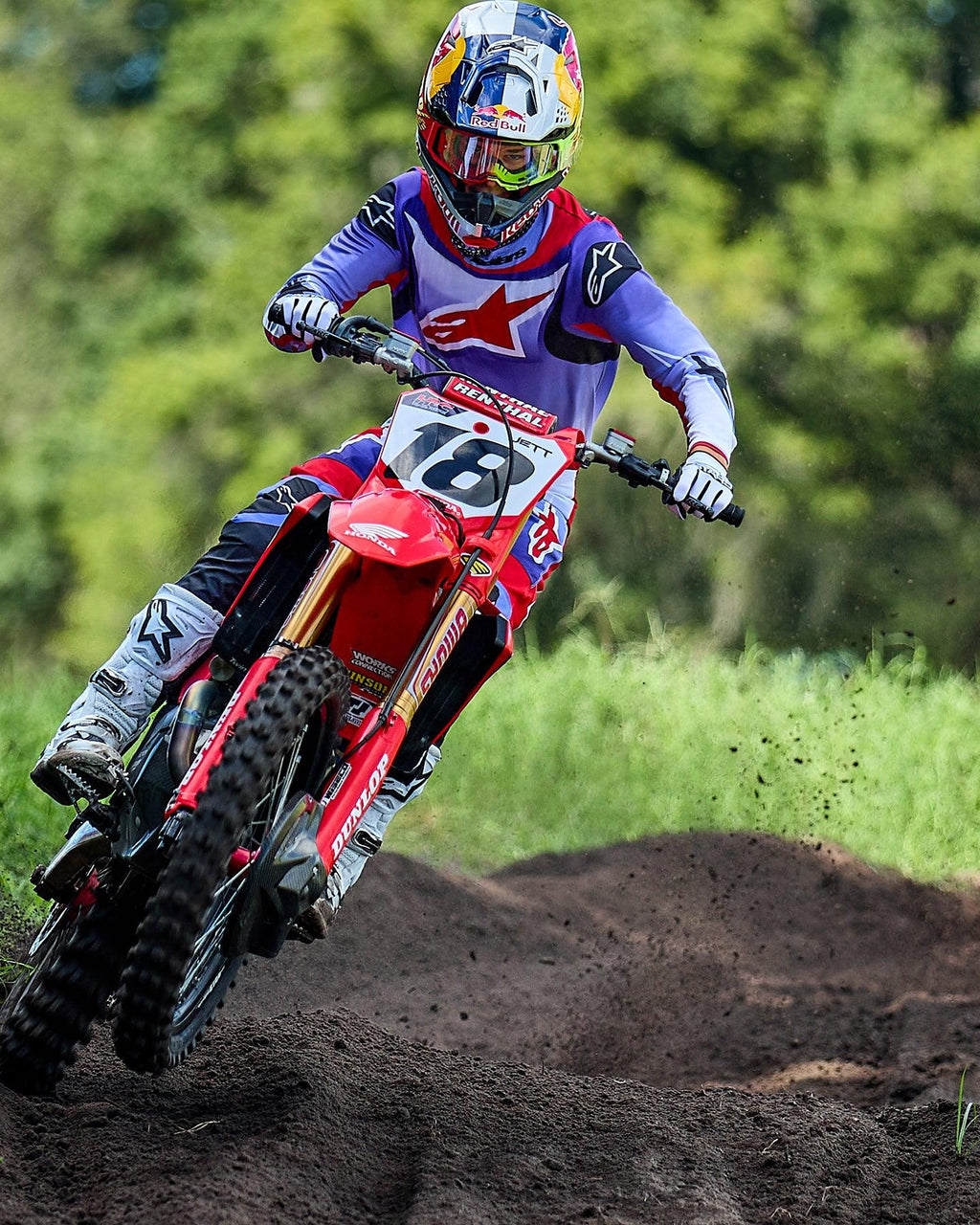 Pilota di motocross su una moto Honda rossa, indossa la maglia Alpinestars 2025 Fluid Wurx viola e rosso brillante, pantaloni coordinati, stivali bianchi e casco Red Bull, mentre affronta una curva su pista sterrata.