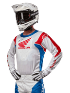 Pilota di motocross con maglia Honda Racer Iconic in bianco, blu brillante e rosso acceso, con un grande logo Honda sul petto, abbinata a un casco Alpinestars bianco e guanti bianchi e neri coordinati, equipaggiamento per prestazioni fuoristrada