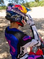 Pilota di motocross Hunter Lawrence con maschera Limited Edition HUNTA96 XXV Supertech Vision, fascia viola con scritta 'H96' e cuciture gialle, casco blu Red Bull, maglia viola e bianca con dettagli neri, guanti bianchi, intento a regolare la maschera in un contesto fuoristrada sabbioso