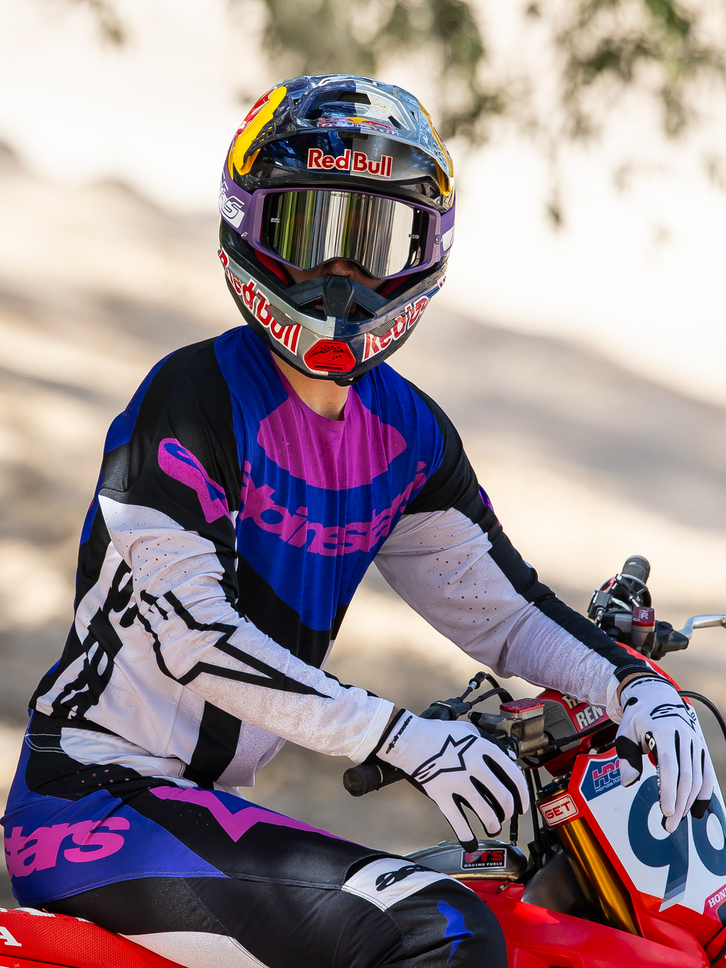 Pilota di motocross su una moto da cross rossa, maschera Alpinestars HUNTA96 XXV Supertech Vision Limited Edition in viola e bianco con lenti a specchio argentate, maglia e pantaloni coordinati viola, nero e bianco, casco Red Bull, guanti bianchi, abbigliamento professionale da motocross
