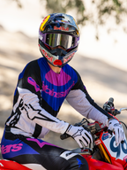 Pilota di motocross su una moto da cross rossa, maschera Alpinestars HUNTA96 XXV Supertech Vision Limited Edition in viola e bianco con lenti a specchio argentate, maglia e pantaloni coordinati viola, nero e bianco, casco Red Bull, guanti bianchi, abbigliamento professionale da motocross